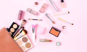 A variety of beauty and cosmetic products are spilling out of a brown bag onto a pink surface.