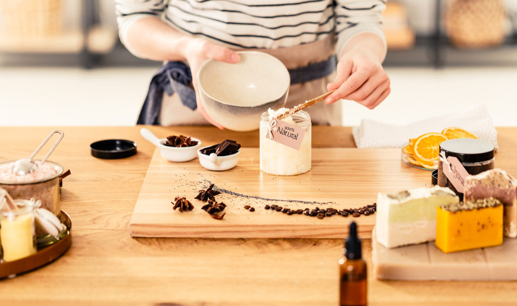 A woman pours ingredients into a jar of homemade beauty products with the label showing the product is 100% organic.