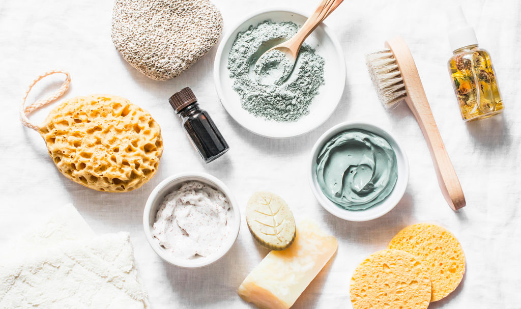A variety of skincare, like face masks, bath salts, and sponges, lay arranged on a white background.