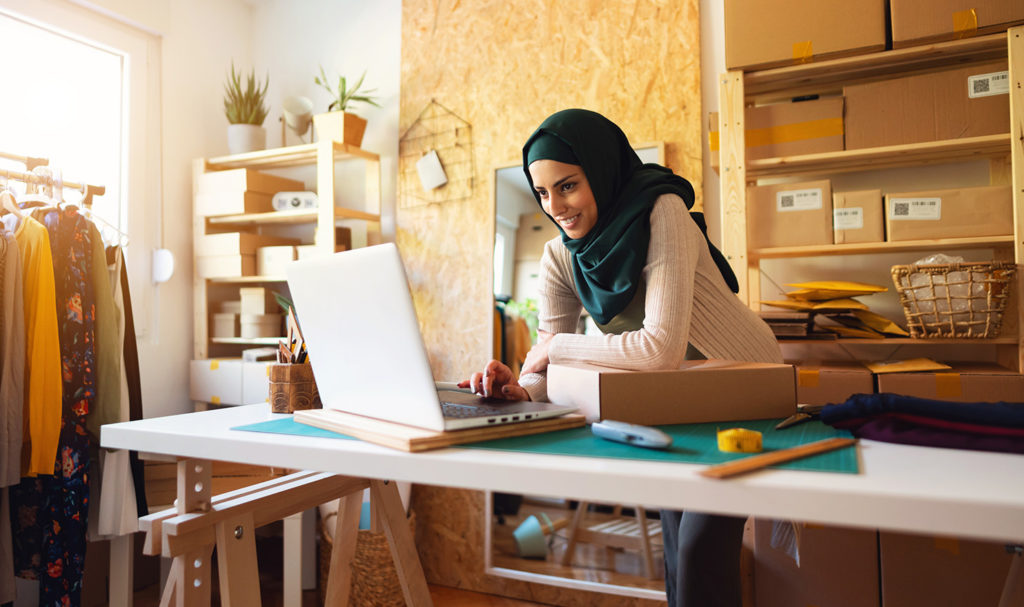 A young woman is in her home office surrounded by boxed products for her business as she leans on a desk to look on her laptop for product liability insurance.