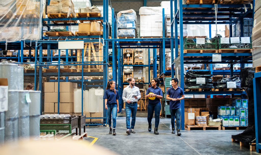 A group of people are walking through a warehouse full of boxed products discussing a new partnership and their new product liability insurance requirements.