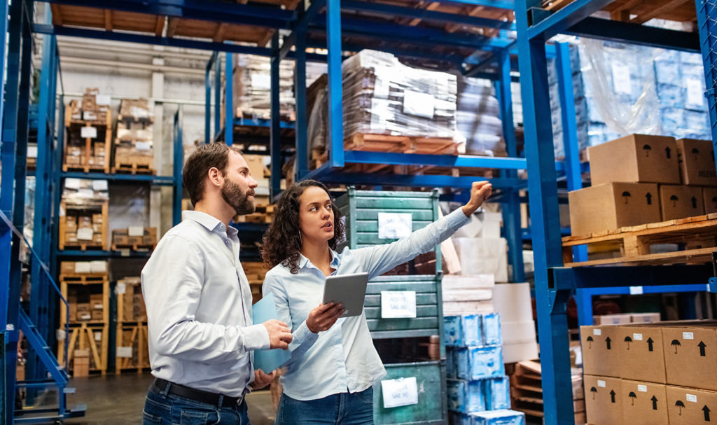 A man and a woman walk through a warehouse with an iPad discussion product liability insurance amongst the shelves of boxes and products.