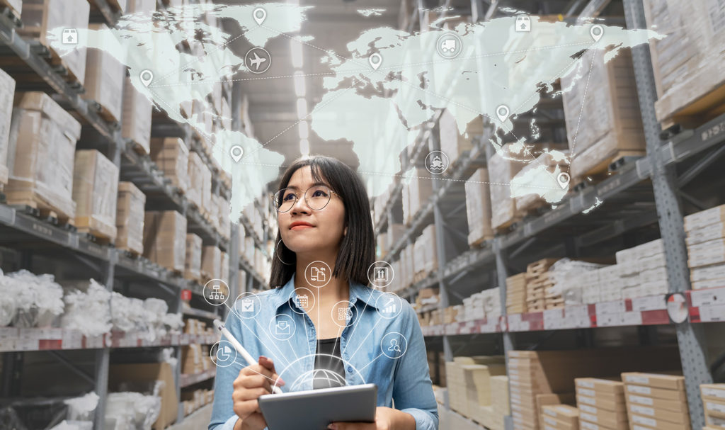 A woman is walking down an aisle of boxes on shelves in a warehouse while holding a tablet and a digital map of the world is over her head.