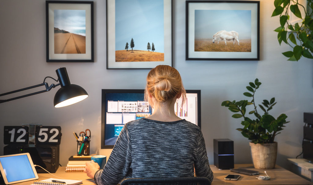 The back of a young woman working from her desk on her small business in her home office surrounding by photos of nature on her wall.