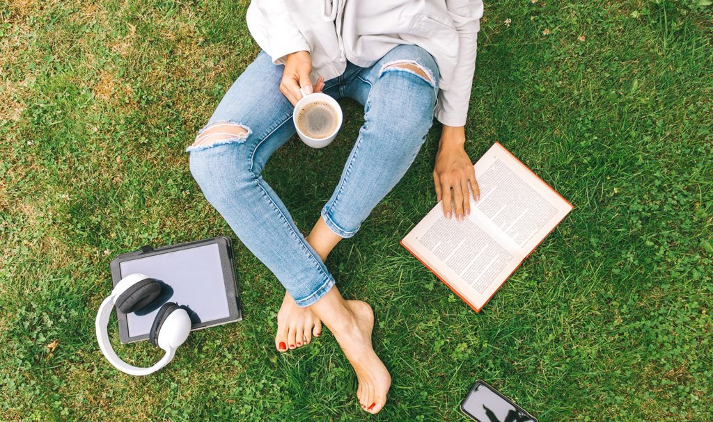 An above view of a person sitting in grass reading a book and holding a coffee next to a tablet, headphones, and phone.