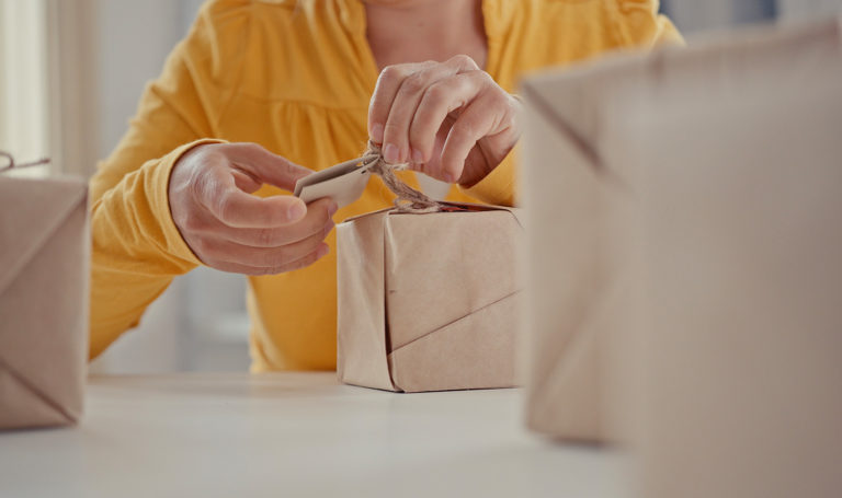 A person in a yellow shirt is packaging up their handmade products into brown paper boxes they have insured with product liability insurance.
