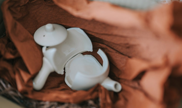 A white porcelain teapot sits broken inside of a dark amber colored tissue paper in a box.