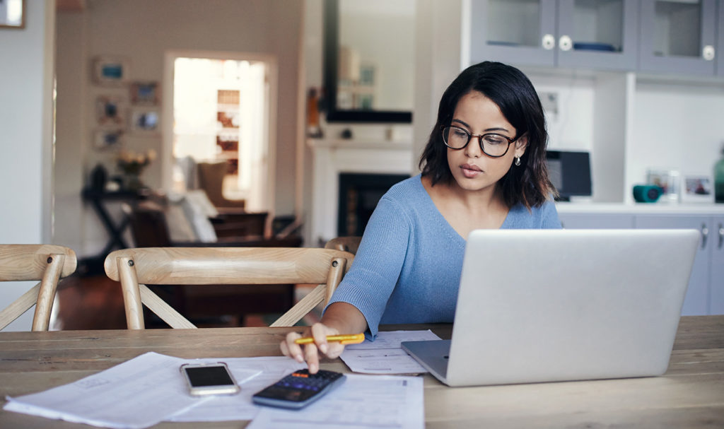 A woman is in her home sitting in her kitchen at the table with a laptop, paperwork, and a calculator as she calculates her risks.