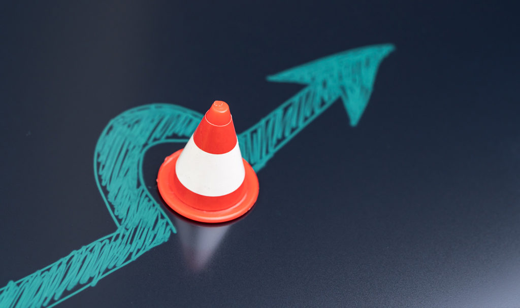 A blue arrow is drawn on a dark service in light blue ink. The arrow is going straight, but has a curve in it as it goes around an orange traffic cone in the way.