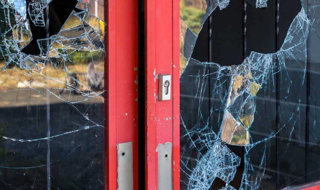 A red door leading inside to a business has glass that has been smashed in, indicating a theft on the business.