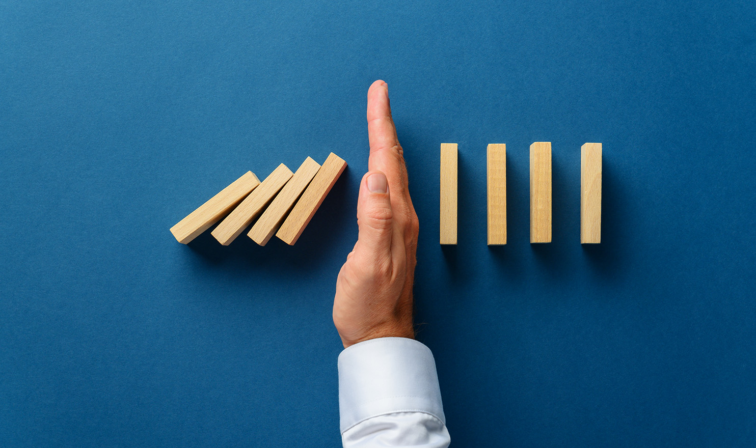 A man's hand in a white professional shirt is stopping a stack of dominoes from falling into the rest of the stack. The image is set on a solid navy blue background.