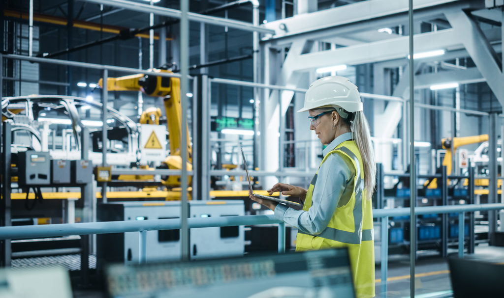 A woman is standing in a manufacturing plant wearing a hard hat and safety vest as she holds a clipboard and is observing the building of cars by automated machinery.