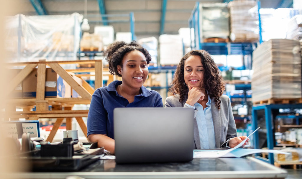 A woman and her advisor smile as they chat at a computer in a warehouse discussing product liability insurance for a business.