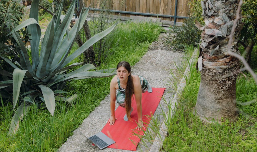 A yoga teacher practices outside on her yoga mat next to her laptop as she works on a virtual yoga teacher certification program.