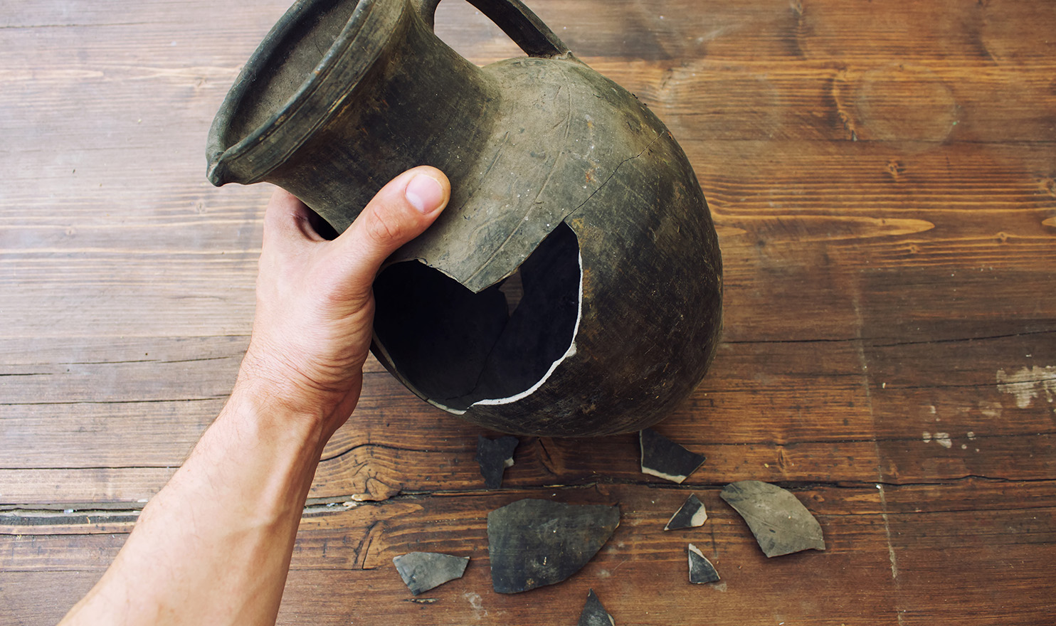 A man's hand is holding a broken vase on a brown wooden table next to the shattered pieces. If he cut his hand on the faulty vase, product liability insurance could help pay his medical bills.