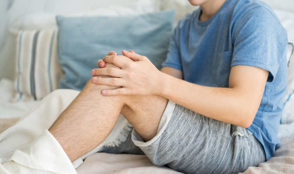 A person is holding their knee while sitting on their bed after injuring themselves on their bed frame. Accidents like this may be covered by product liability insurance.