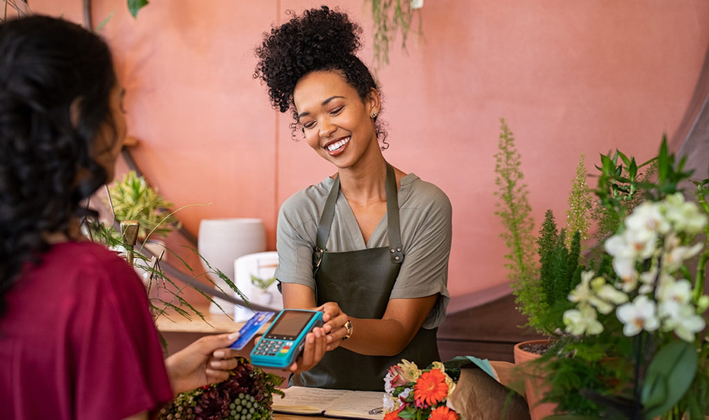 A small business owner is standing behind the counter in her shop as she helps a customer complete a transaction on a payment device.
