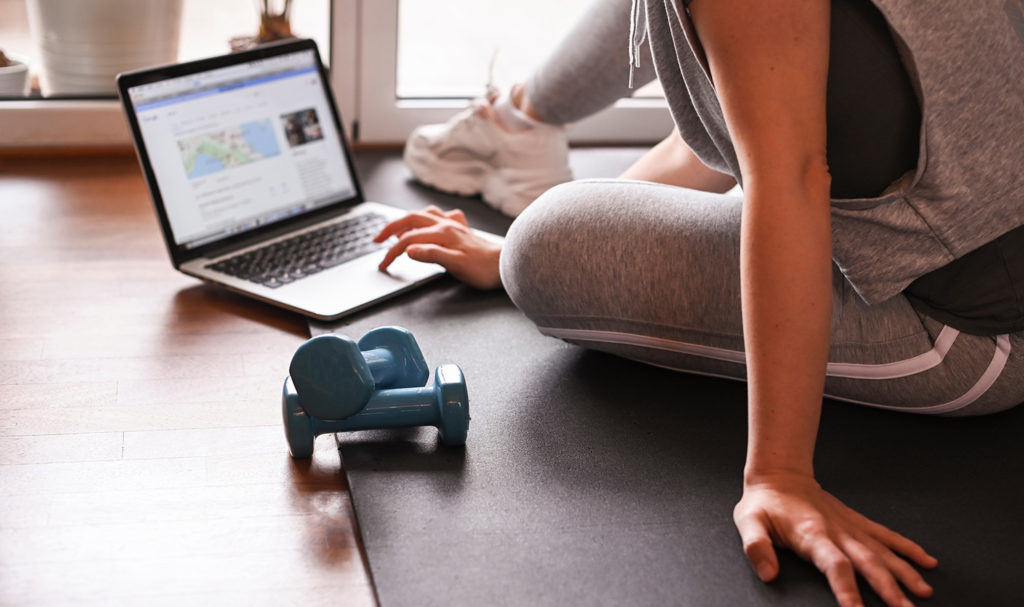 A woman is sitting on a yoga mat on the floor next to weights while she works on making her online personal training business plan on her laptop.