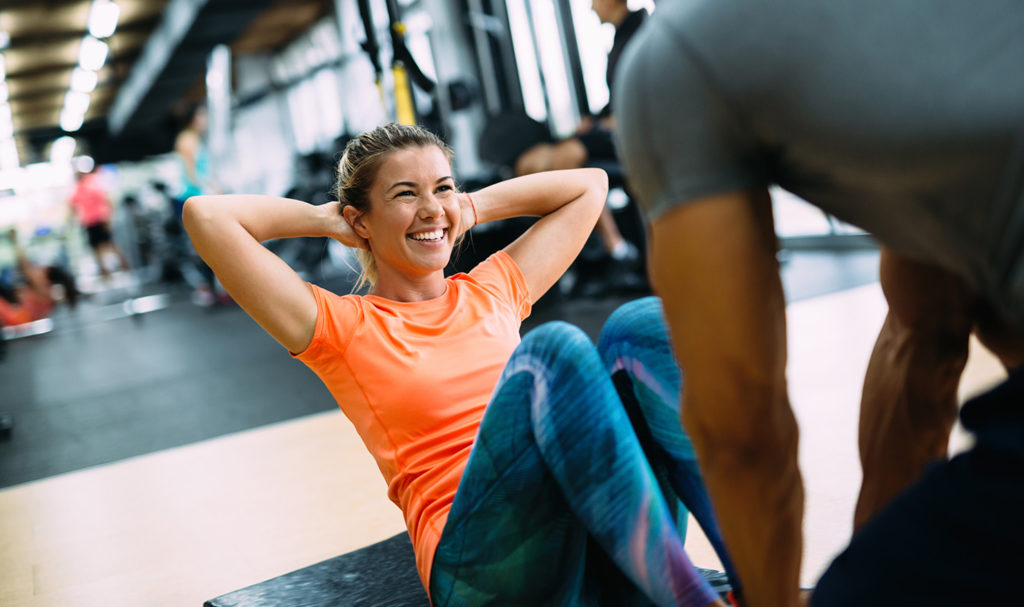 A personal trainer is helping his client with sit-ups in a gym. He is able to find clients to work with because he took the time to make a business plan for personal trainers.