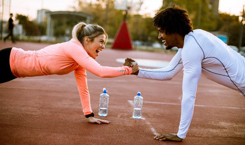 A personal trainer and their client are high fiving each other from a push up position on the ground of an outdoor workout space. Making a business plan for personal trainer businesses helps you feel more confident as a trainer and find the right clients to work with.