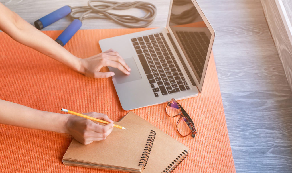 An above view of someone working on a laptop on a yoga mat next to some workout equipment. They are writing on a notepad as they work on planning their finances and operations as part of their business plan for a personal trainer.