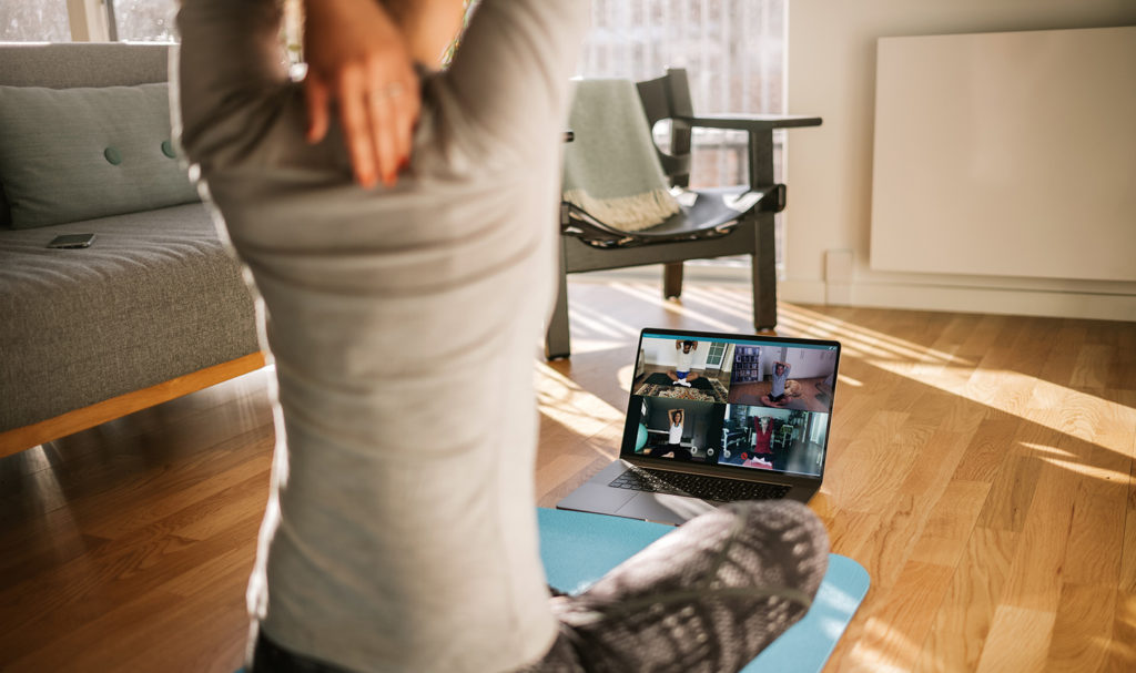 A personal trainer is sitting on a yoga mat in her home in front of her laptop as she walks a group of clients on a virtual call through some stretches.