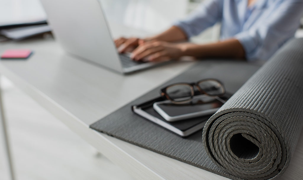 A personal trainer is working on a business plan on their laptop with their workout equipment, glasses, and notebook sit beside them.