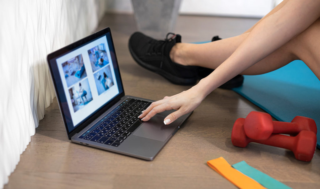 A person is sitting on a blue yoga mat on the floor of their home next to some weights as they work on their laptop to complete a personal trainer certification course.