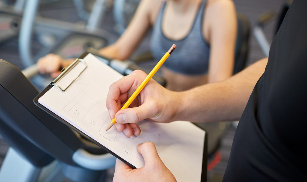 A personal trainer is taking notes on a clipboard while working with their client. Fitness business names can help trainers find clients easier.