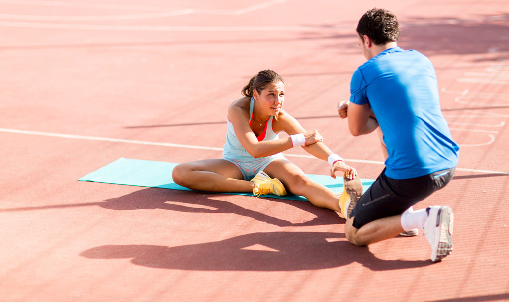 An athletic training is helping a client work on their stretching while they sit outdoors on a track. After researching personal trainer business names, the trainer could pick a good name for their business and start getting clients.