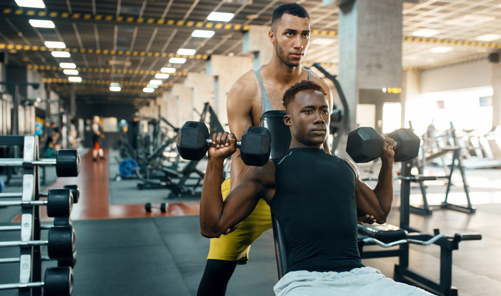A personal trainer is helping his client learn strength training by guiding him through lifting weights in the gym. He brainstormed some personal training name ideas with some clients to get a feel for which one they like the best and would identify him by.