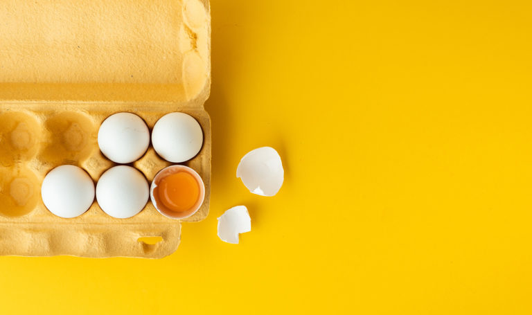 A carton of eggs sits on a yellow background, and one egg is broken in the carton with the shell pieces lying next to it. Food manufacturing insurance may help if a bad egg gets a customer sick.