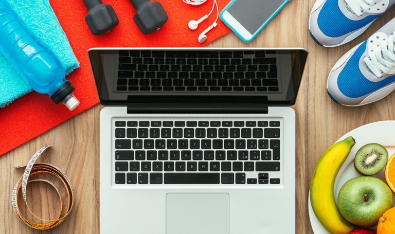 A laptop on a table sits beside weights, running shoes, water bottle, fruits, and various workout equipments.
