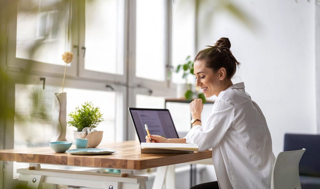 A young woman sits at a desk in a home office and writes notes down on a pad of paper. She is surrounded by a laptop, plants, and other working tools as she makes getting product liability insurance part of her business plan.