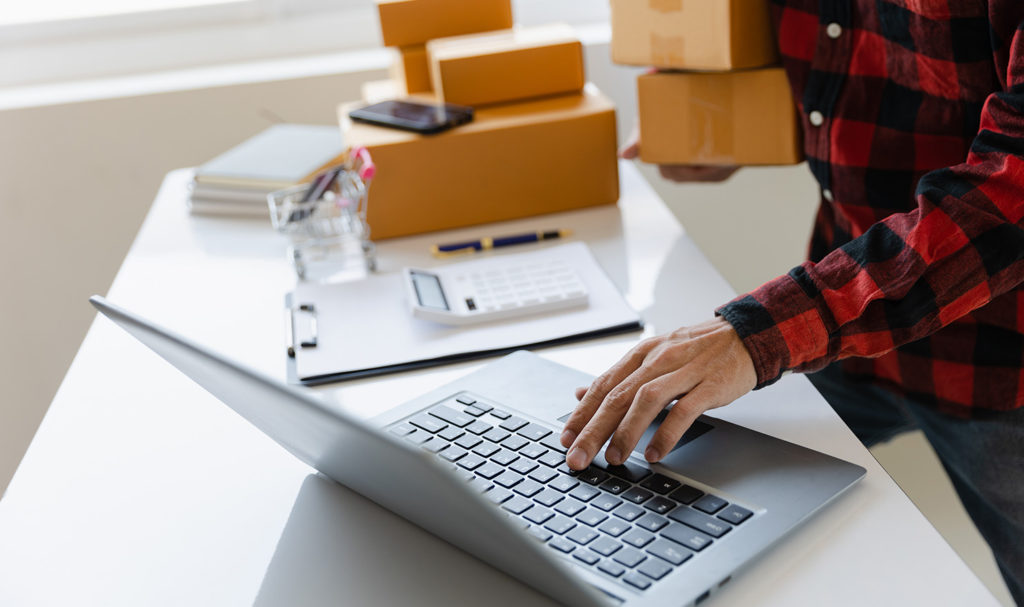 A business owner's hands are working on a laptop on a table next to products ready in packages to be shipped. Product insurance for small business can help to protect them from claims that arise from these products.