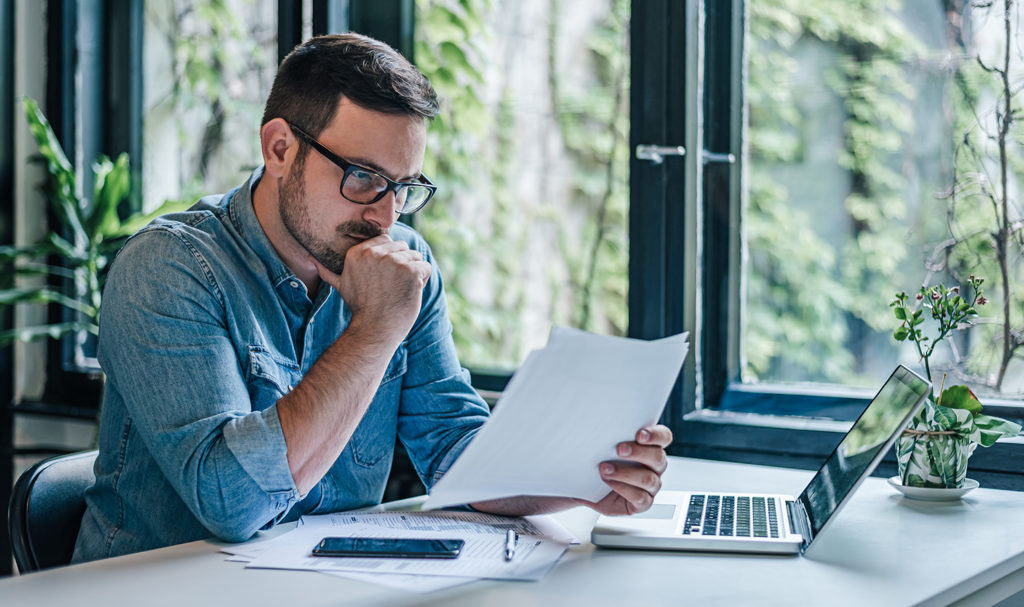 A young business owner is looking over paperwork in his home office next to his laptop as he decides on what product liability insurance to buy.