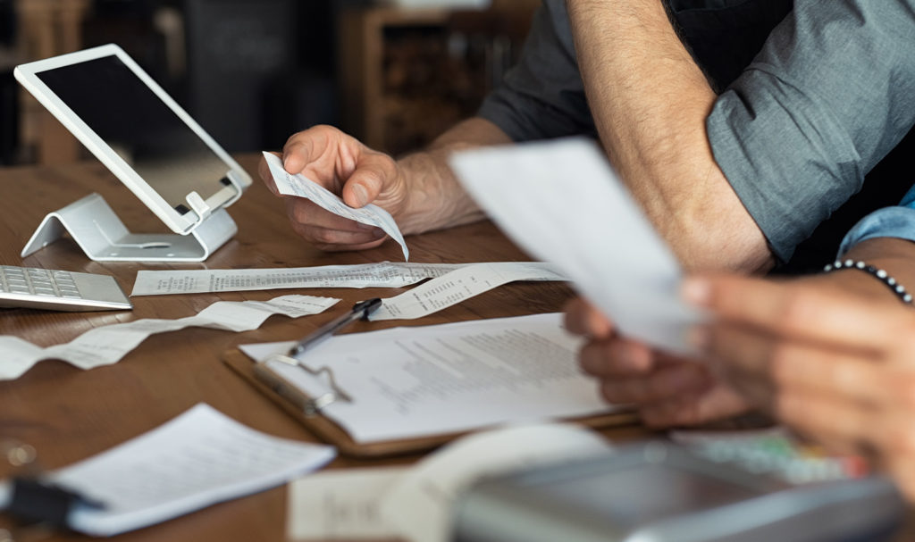 The hands of two business owners are sorting through paperwork and receipts as they work on a tablet on their small business plan. Finding the cheapest product liability insurance is a top priority for them and their business finances.