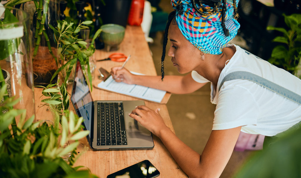 A young woman is working in her small business floral shop as she leans down to work on a laptop on a desk. Getting small business product liability insurance will aid her in her business planning process.