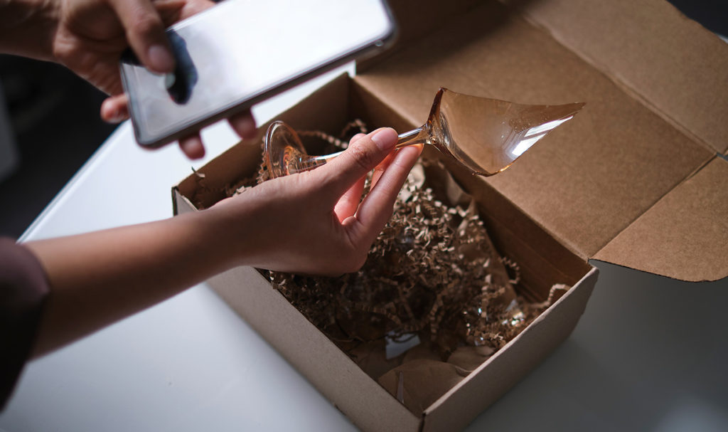 A photo of a person holding a broken glass over the top of a package, while holding their phone in the other hand to document the product damage.