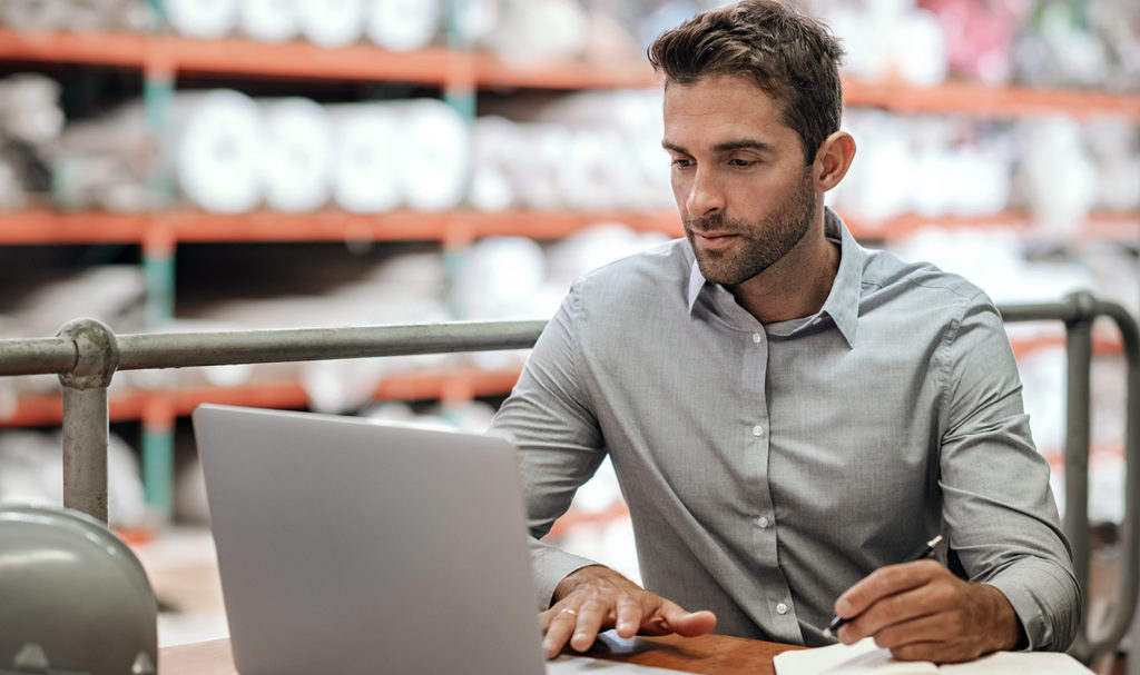A man in a button up shirt works on a laptop in a warehouse as he looks at the best product liability insurance options for his business.