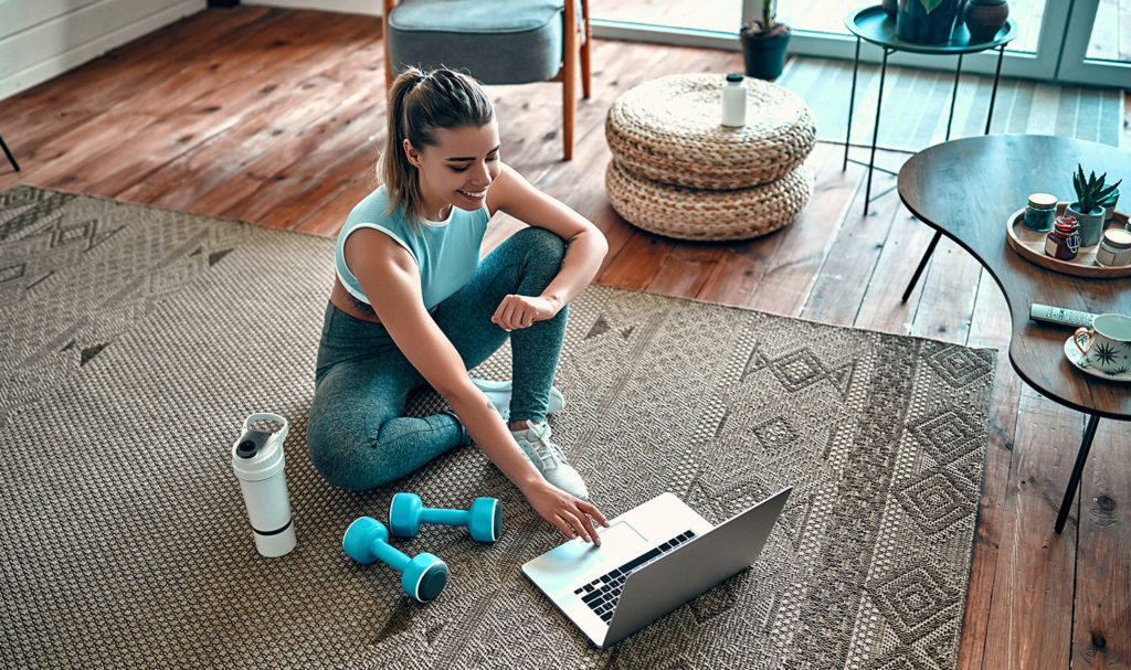 A personal trainer sits on the floor of her home while she works on fitness marketing strategies on her laptop next to her workout gear.