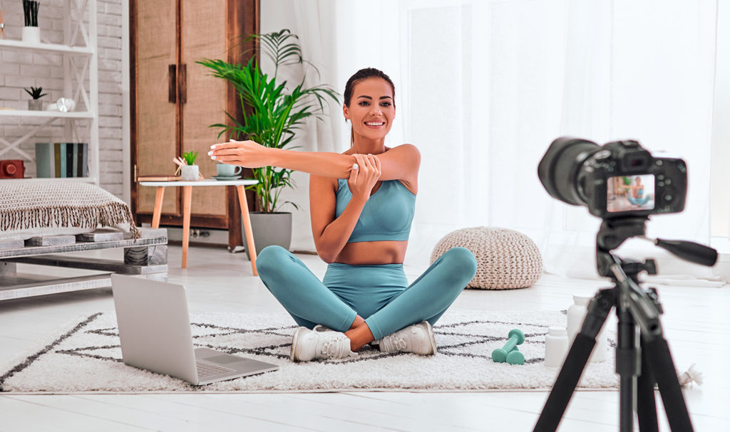 A personal trainer is sitting on the floor of her home in front of a camera mounted to a tripod so she can record a workout. Her laptop is next to her where she was working on her personal trainer marketing plan.