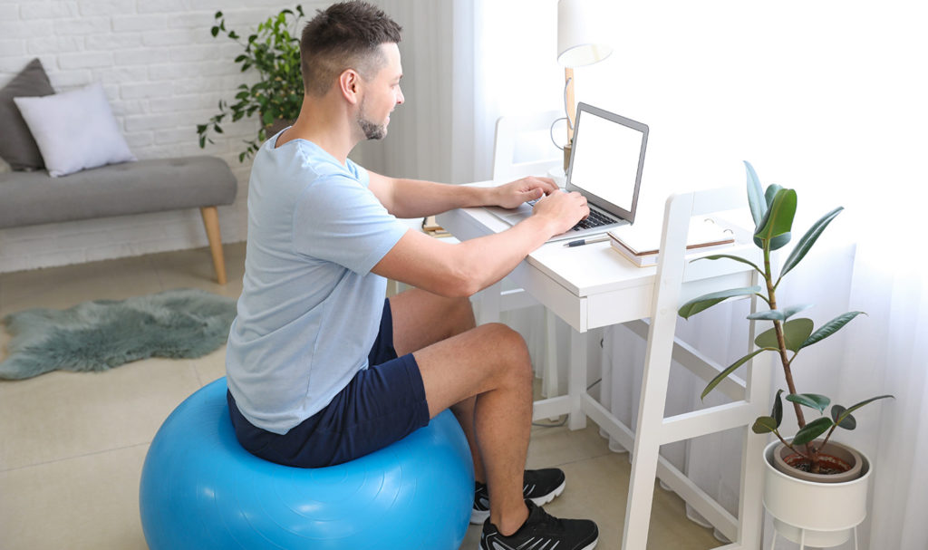 A personal trainer sits on a blue yoga ball inside his home office as he works at a desk on his laptop to create facebook ads for personal trainers so he can better promote his business online.