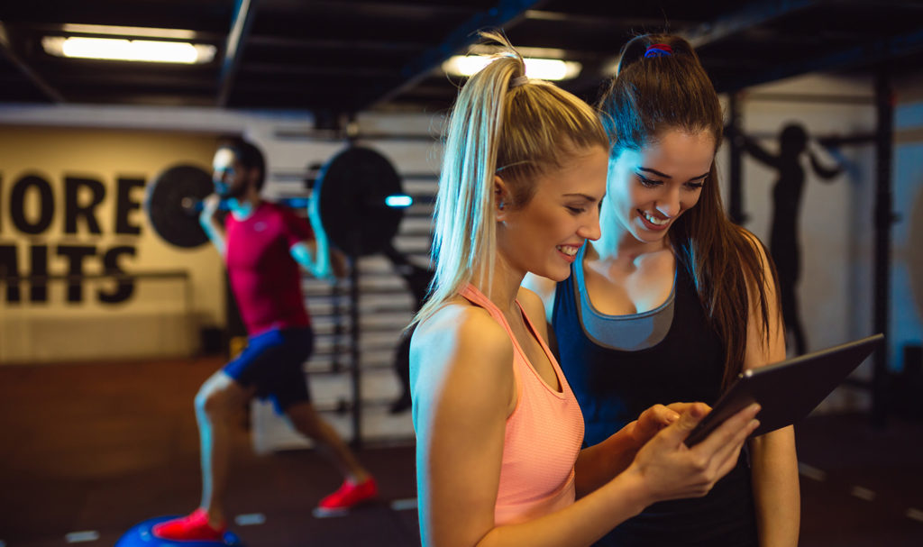 A personal trainer is talking with her client and showing her a form on her ipad. Her client can fill this out to leave a testimonial of her experience working with the personal trainer. The trainer can use testimonials to increase their personal trainer marketing strategies.