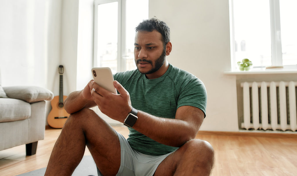 A personal trainer is sitting on the ground of their home looking at their recent marketing data on their smart phone. This is a great way to make sure your fitness marketing strategies are working.