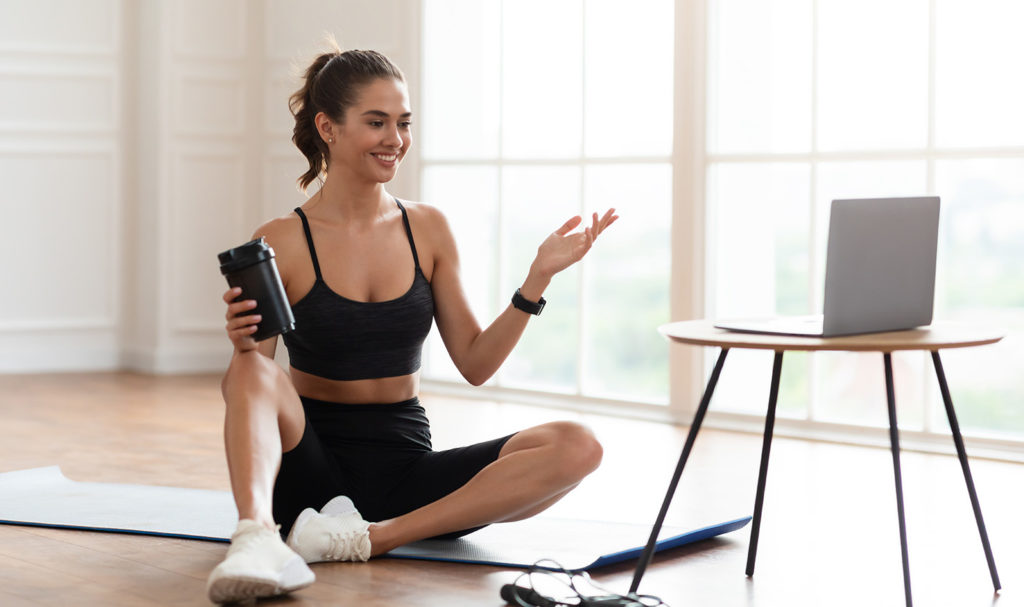 A woman on a yoga mat talks towards her laptop. She is recording a virtual training session to help her learn how to get clients as a personal trainer.