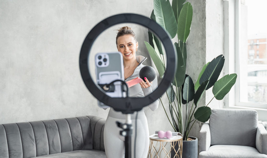 A personal trainer is standing in front of a ring light with a phone mounted to it so she can record herself doing a workout. Creating online videos is a great way to up your marketing fitness game.