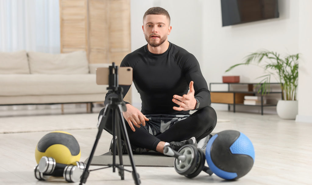 A man on a yoga mat with weights and medicine balls speaks to a camera phone on a tripod after learning how to sell fitness programs online.