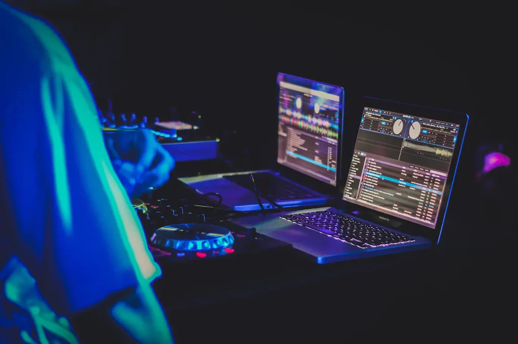 Two laptops are open in a dark room showing DJ software while the DJ stands out of focus to the left side of the frame.