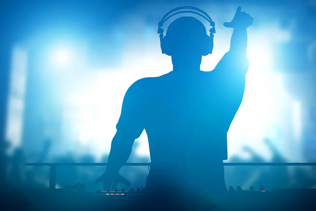 Image of a DJ standing on stage with their back to the camera and arms outstretched in front of their equipment and a crowd.